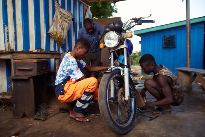 Teye sitter på huk ved siden av en motorsykkel, to andre hjelper til. Foto: Henry KD