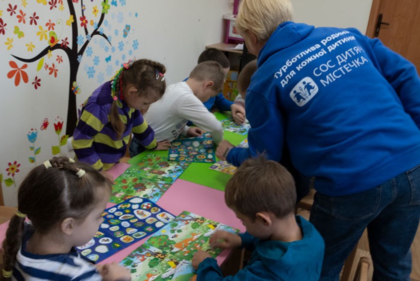 En sosialarbeider på et senter som tilbyr oppfølging, støtte, lek og læring for barn og familier, hjelper flere barn som sitter rundt et bord med kreative aktiviteter. Hun har lyst halvlangt hår og blå vest med SOS-barnebyers logo på ryggen. Foto: Katerina Ilievska
