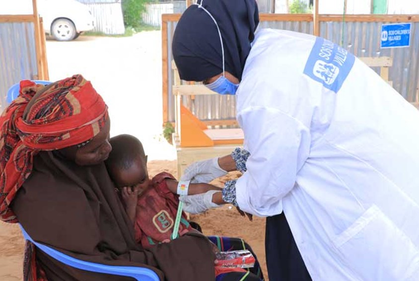 En sykepleier i hvit uniform med SOS-barnebyers logo på ryggen og blå hijab, måler tykkelsen på armen til et lite barn som sitter på morens fang. Foto: Sunshine Media