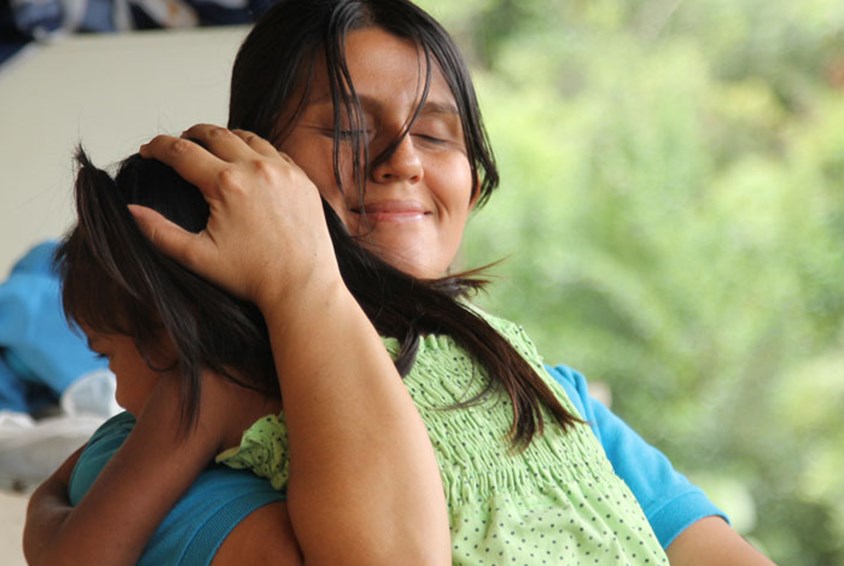 En fostermor i SOS-barnebyer holder ei jente tett inntil seg og klemmer henne. Moren har mørkt langt hår i hestehale og blå t-skjorte, jenta har grønn kjole. Foto: Giti Carli Moen