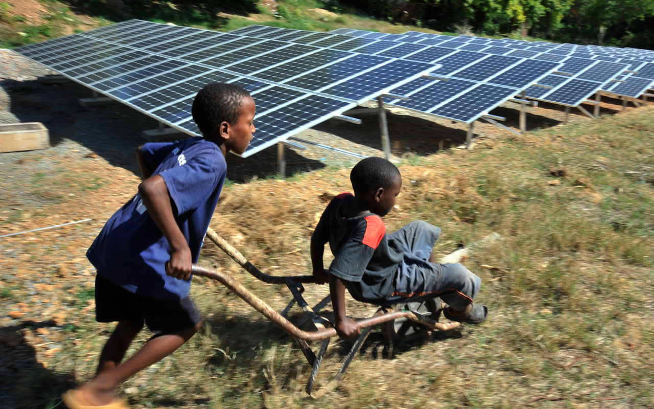 En gutt fra et afrikansk land kjører en annen gutt i ei trillebår. I bakgrunnen er det en rekke med solcellepanel på bakken. Foto GIZ