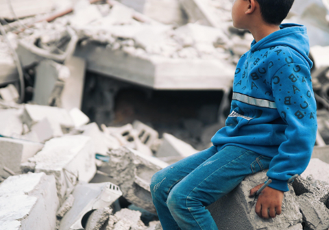 En gutt i blå genser og bukse, sitter i ruinene i Gaza, Palestina. Foto: Hosny Salah