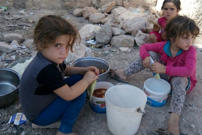 Tre barn på flukt fra krigen i Syria, sitter på bakken blant ruiner og spiser maten de har fått utdelt i bøtter. Foto: SOS-team