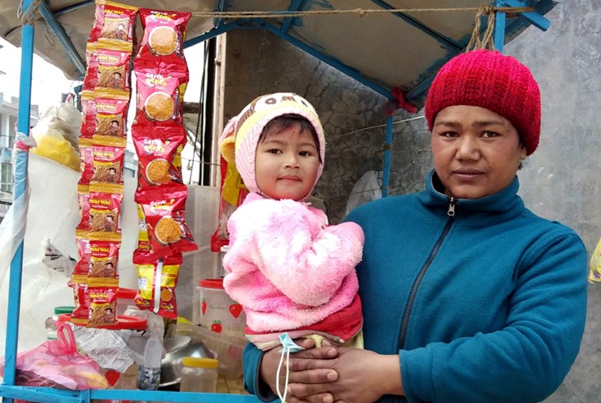Samjhana (38) fra Katmandu i Nepal har datteren (3) på armen. De står foran den lille matboden sin. Datteren er kledd i rosa og Samjhana har blå jakke og rød lue. Foto:  Pearl Sandh