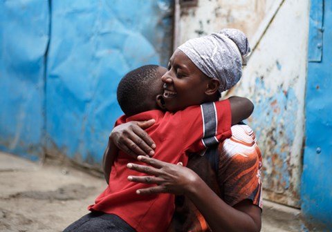 En gutt fra Kenya i rød og blå t-skjorte gir moren sin en god klem. Hun har lyselilla tørkle på hode og mønstret, oransje kjole. Foto: Jakob Fuhr