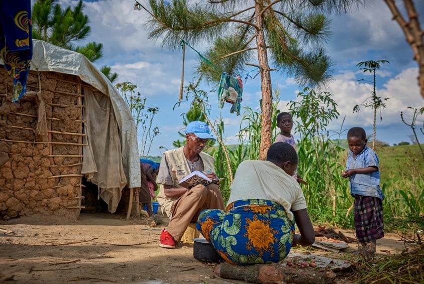 SOS-barnebyer driver flere forebyggende familieprogram i Uganda. Foto: Joris Lugtigheid SOS-barnebyer driver flere forebyggende familieprogram i Uganda. En mor sitter på bakken og skreller grønnsaker, hun har blomstrete skjørt og lys t-skjorte, to av barna hennes står og ser på. SOS-barnebyers medarbeider sitter ved siden av. Han har blå caps, beige vest med SOS-barnebyers logo, bun bukse og røde joggesko. I bakgrunnen er huset de bor i, med vegger laget av leire og et klede foran døra. Foto: Joris Lugtigheid