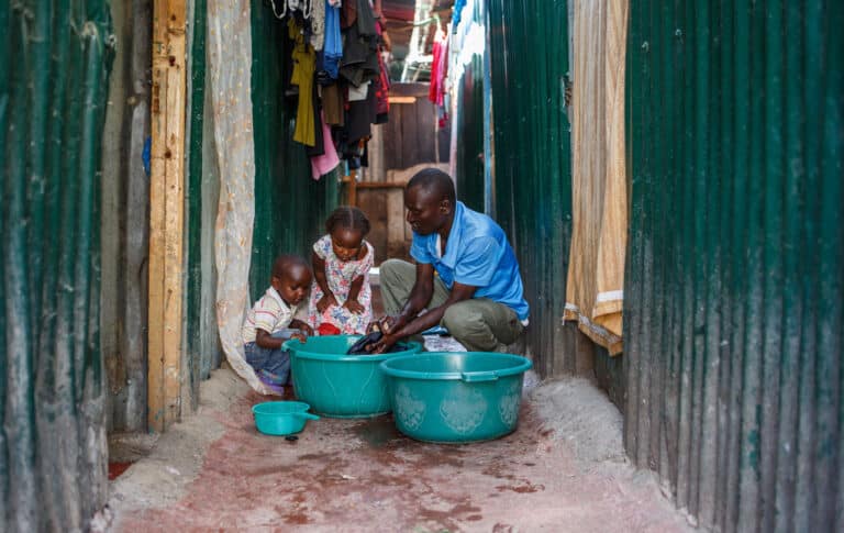 Anthony, i blå skjorte og beige bukse, vasker klær i to turkise baljer. Sønnen står ved siden av og i stripete t-skjorte med krage og knapper, datteren som står i midten har blomstrete kjole. Foto: Jacob Fuhr