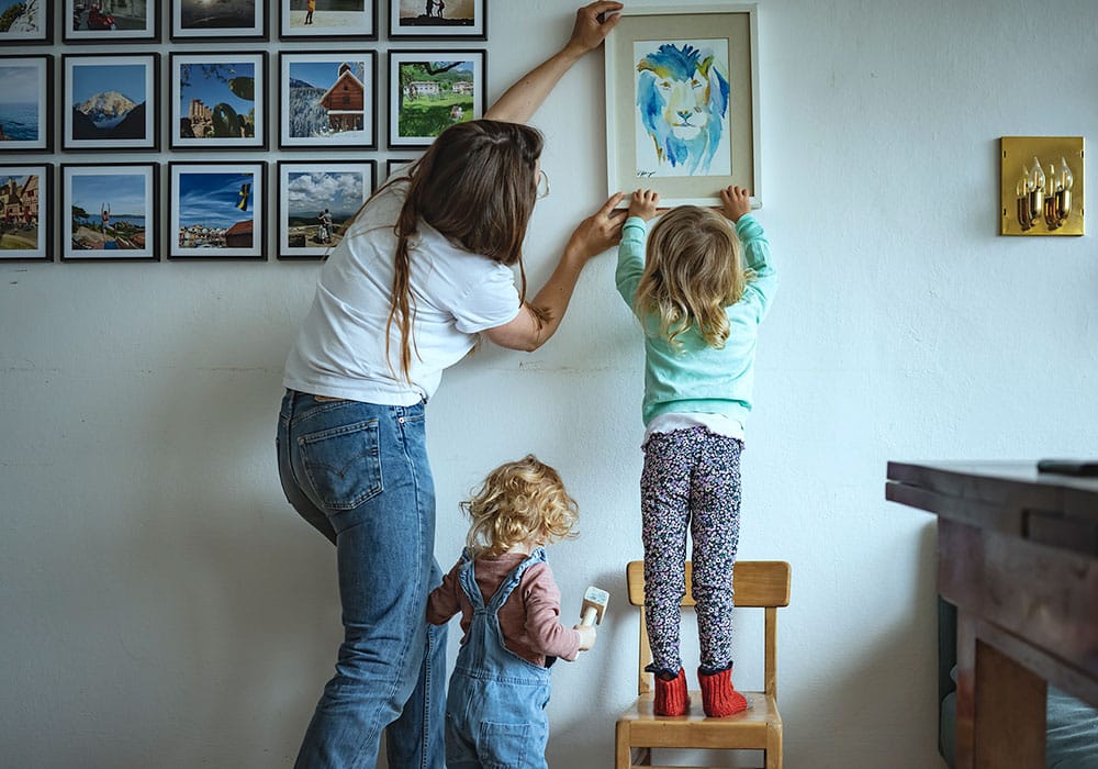 En mor og to barn, alle med ryggen til henger opp en innrammet tegning på veggen. Moren har mørkeblondt langt hår, hvit t-skjorte og jeans, den minste datteren har lyst krøllet hår, rosa genser og snekkerbukse, hun holder en hammer i hånda. Den eldste har langt bølgete hår, hun har grønn genser og mønstrete bukse, og står på en liten barnestol. Foto: Gerhard Berger