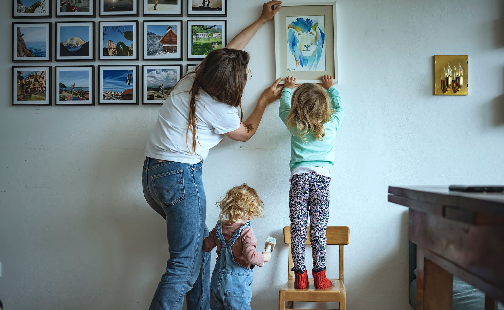 En mor og to barn, alle med ryggen til henger opp en innrammet tegning på veggen. Moren har mørkeblondt langt hår, hvit t-skjorte og jeans, den minste datteren har lyst krøllet hår, rosa genser og snekkerbukse, hun holder en hammer i hånda. Den eldste har langt bølgete hår, hun har grønn genser og mønstrete bukse, og står på en liten barnestol. Foto: Gerhard Berger