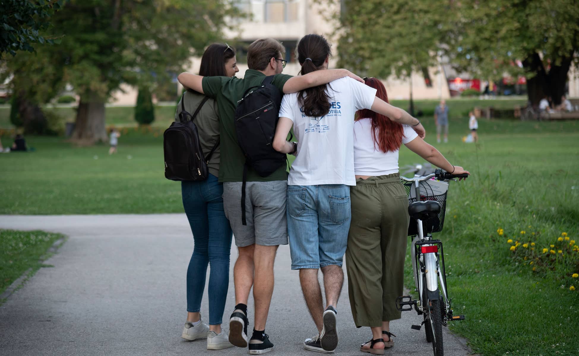 Unikt samarbeid: To unge jenter og to unge gutter går på en vei i en park. De holder hverandre rundt skulderen, den ene triller en sykkel. Bildet er tatt bakfra. Foto: Katerina Ilievska