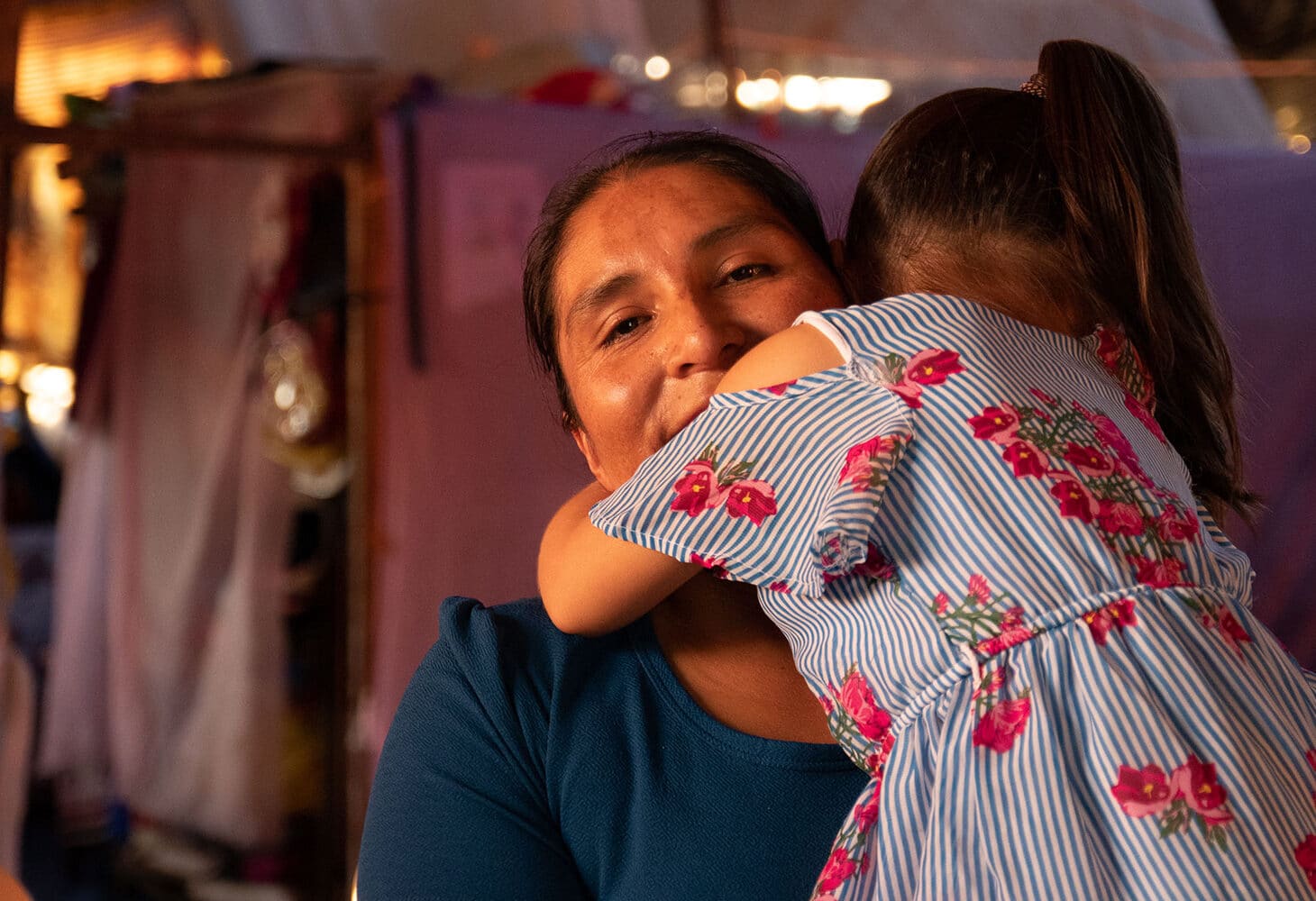 Bolivia: Hilda, med mørkt hår i hestehale og blå genser, holder den yngste datteren på armen. Hun har ryggen til, mørkt hår i hestehale og hvit og blå stripte kjole med rød blomster. Foto Monica Garcia