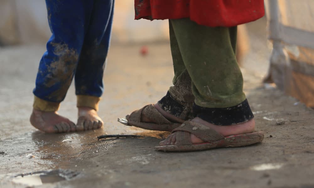 Bilde av barneføtter som står på en sølete vei. Ett barn er barbeint og har søle langt oppover den blå joggebuksa, det andre barnet har beige sølete sandaler og grønn bukse. Foto: Ari Jalal