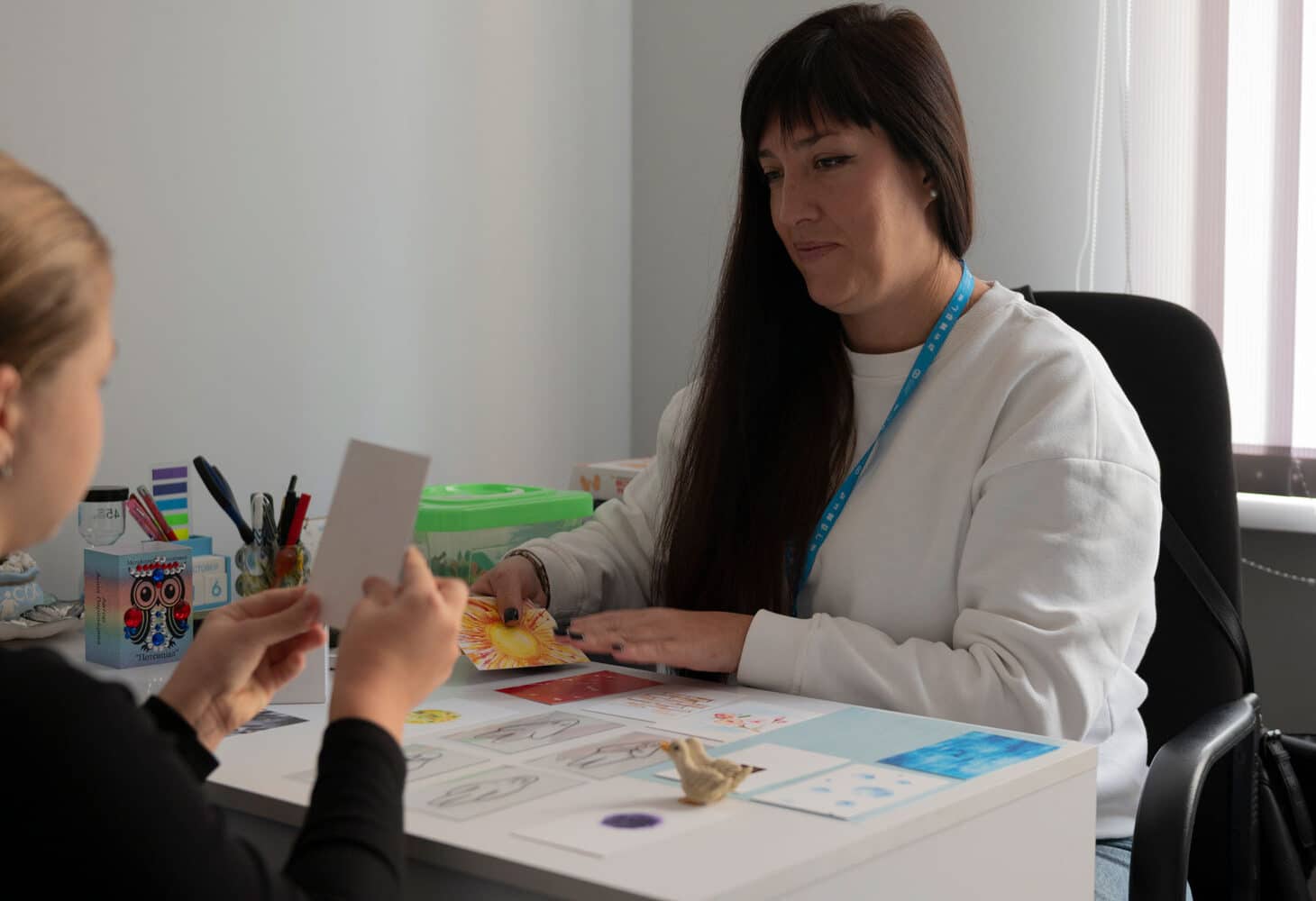Iryna, psykolog i SOS-barnebyer Ukraina. med langt mørkt hår og hvit genser, sitter ved et bord sammen med ei jente. Hun har blondt hår og mørk genser. Foto Katerina Ilievska