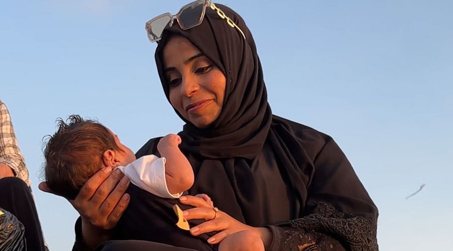 En av SOS-barnebyers medarbeidere i Gaza sitter på bakken med en liten baby i armene. Hun har svart lang kjole og svart hijab. Foto Lama Qattush