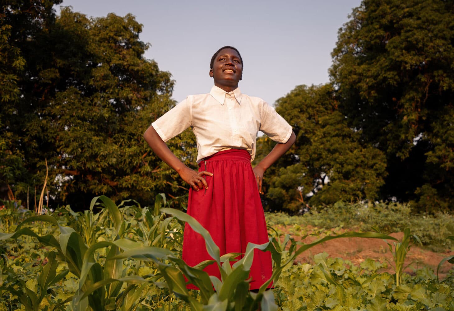 Ung kvinne fra Malawi med kort, mørkt år, hvit kortermet bluse og rødt skjørt står med hendene i siden og skuer utover. Bak henne er det en gresslette og trær. Foto Cornelis van Heerden