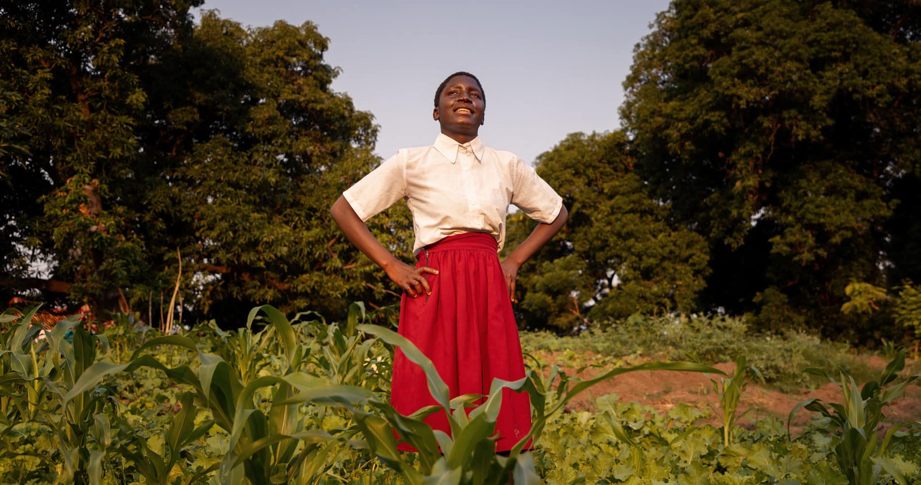 Ung kvinne fra Malawi med kort, mørkt år, hvit kortermet bluse og rødt skjørt står med hendene i siden og skuer utover. Bak henne er det en gresslette og trær. Foto Cornelis van Heerden