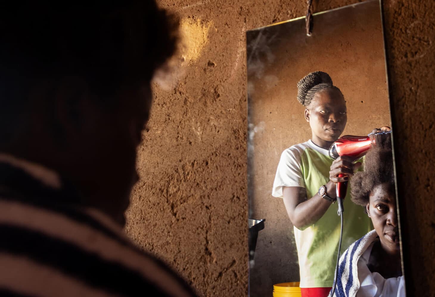 En ung kvinne fra Malawi, med gul t-skjorte og håret i topp, som har utdannet seg til frisør står og føner håret til en kvinnelig kunde. Vi ser begge to i et speil på veggen. Foto Cornel van Heerden