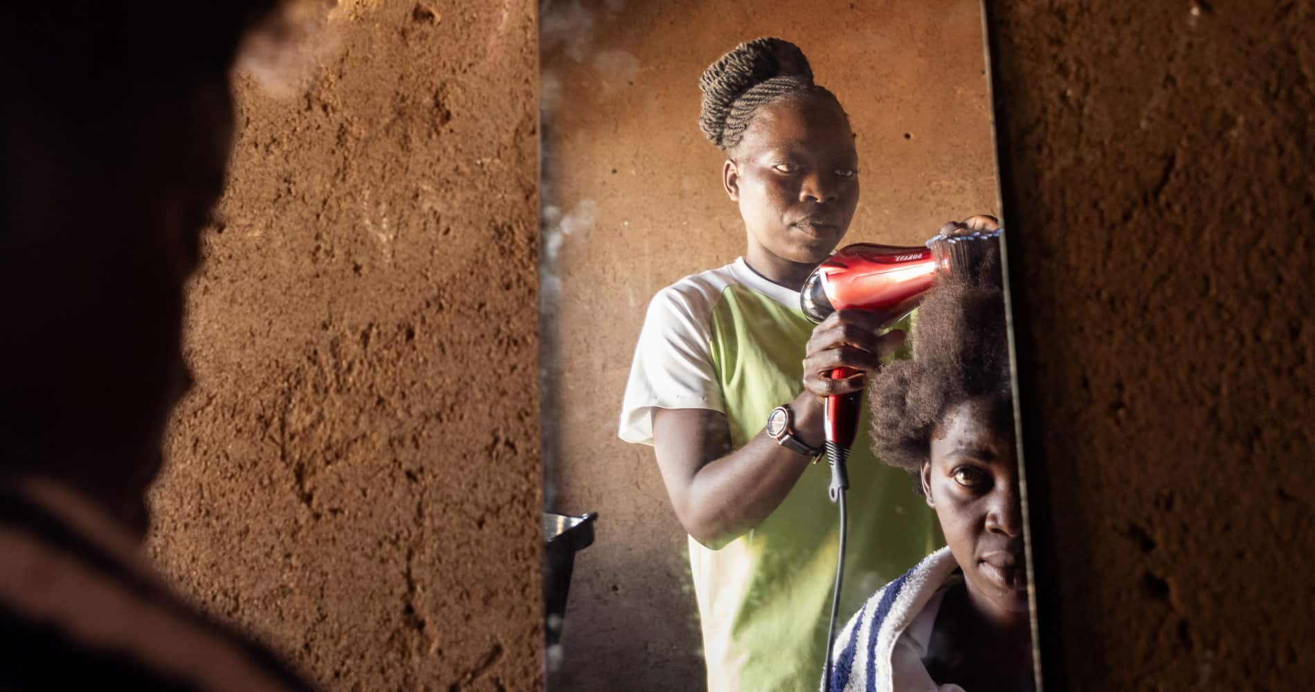 En ung kvinne fra Malawi, med gul t-skjorte og håret i topp, som har utdannet seg til frisør står og føner håret til en kvinnelig kunde. Vi ser begge to i et speil på veggen. Foto Cornel van Heerden
