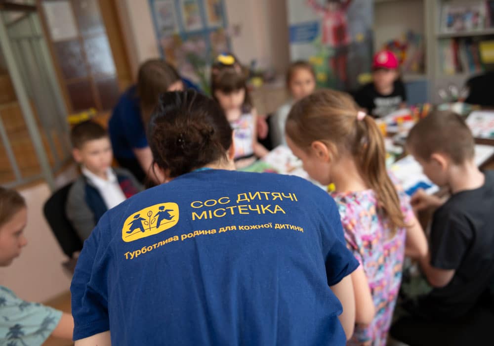 En av SOS-barnebyers medarbeidere i Ukraina sitter med ryggen til. Hun har blå t-skjorte med SOS-barnebyer Ukrainas logo. Rundt bordet sitter barn og jobber med lekser. Foto Katerina Ilievksa