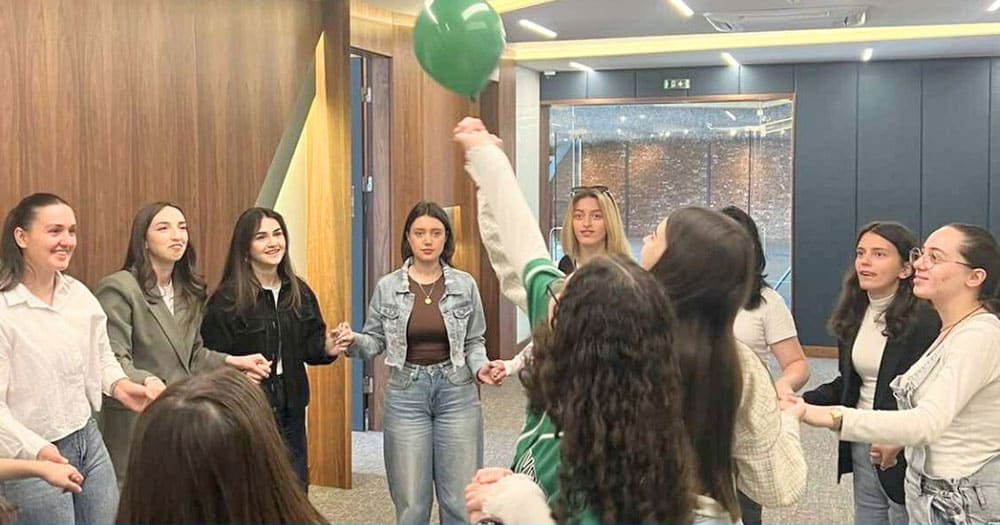 En gruppe unge jenter står i ring og holder hverandre i hendene. De er med på en samarbeidsaktivitet der en ballong skal holdes i lufta. Foto SOS-barnebyer Kosovo