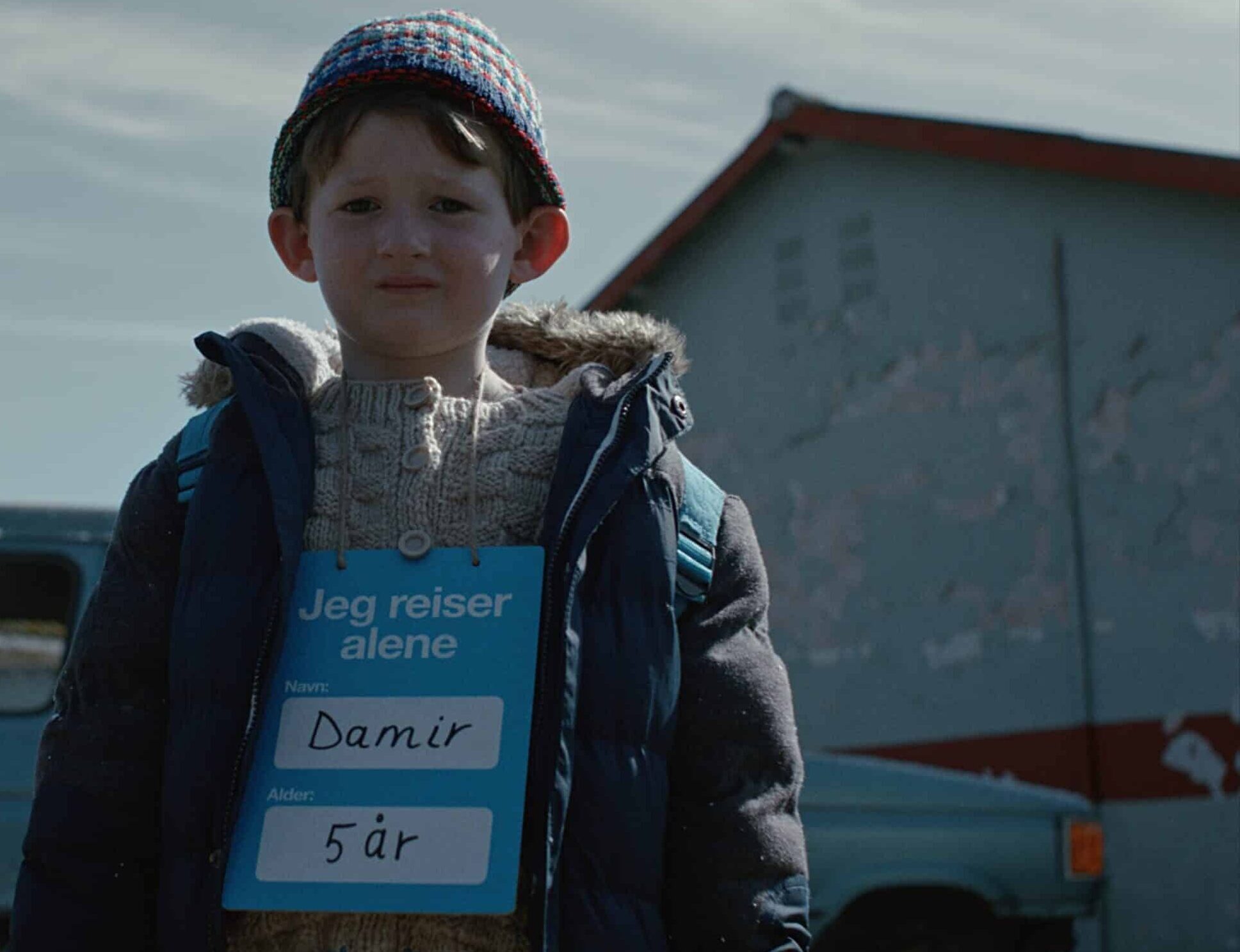 En gutt på fem år står ute foran en ødelagt bil med et skilt rundt halsen der det står «Jeg reiser alene». Bildet er fra SOS-barnebyers kampanjefilm og symboliserer barn som mister tryggheten sin i Ukraina