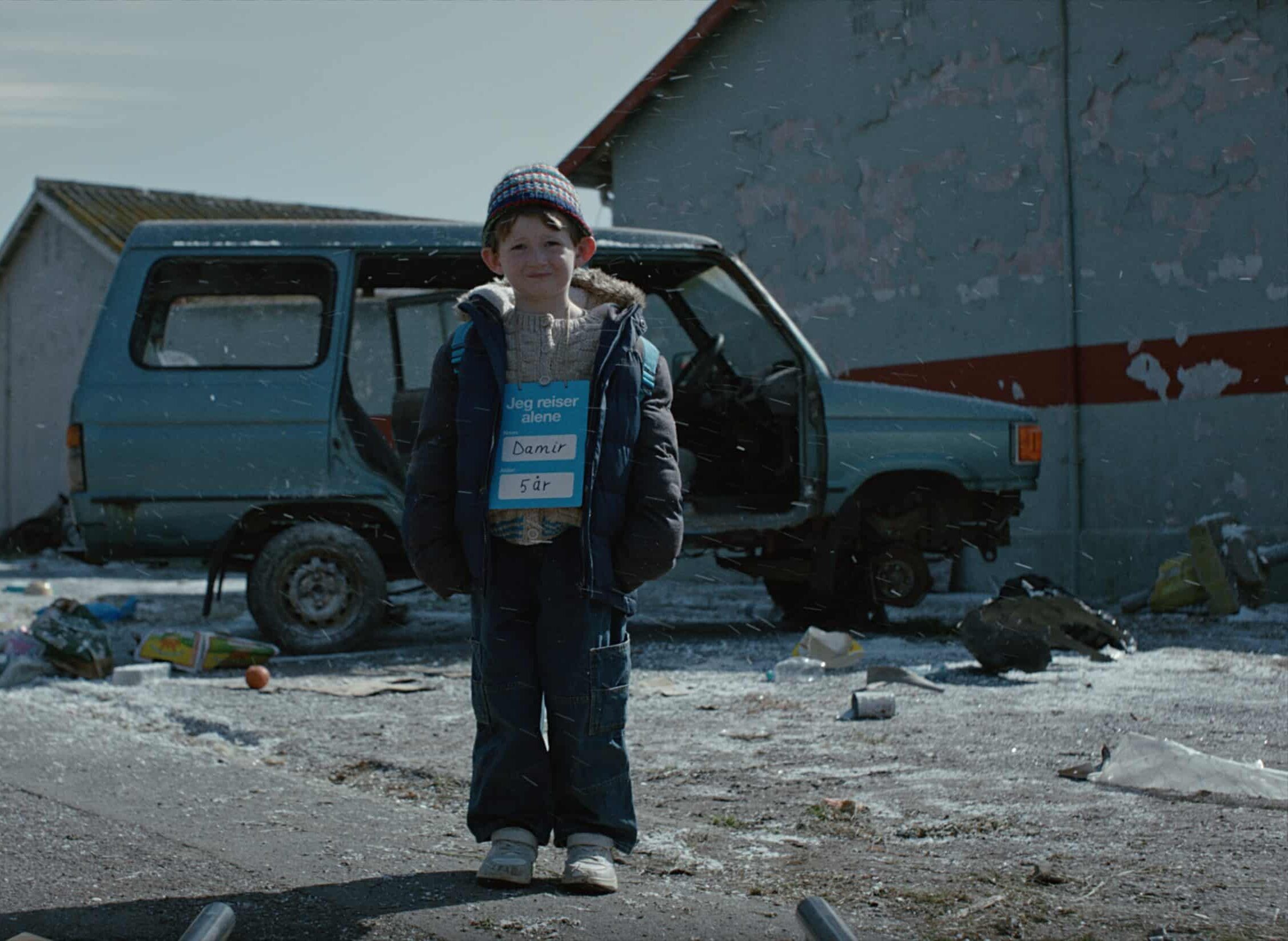 En gutt på fem år står ute foran en ødelagt bil med et skilt rundt halsen der det står «Jeg reiser alene». Bildet er fra SOS-barnebyers kampanjefilm og symboliserer barn som mister tryggheten sin i Ukraina