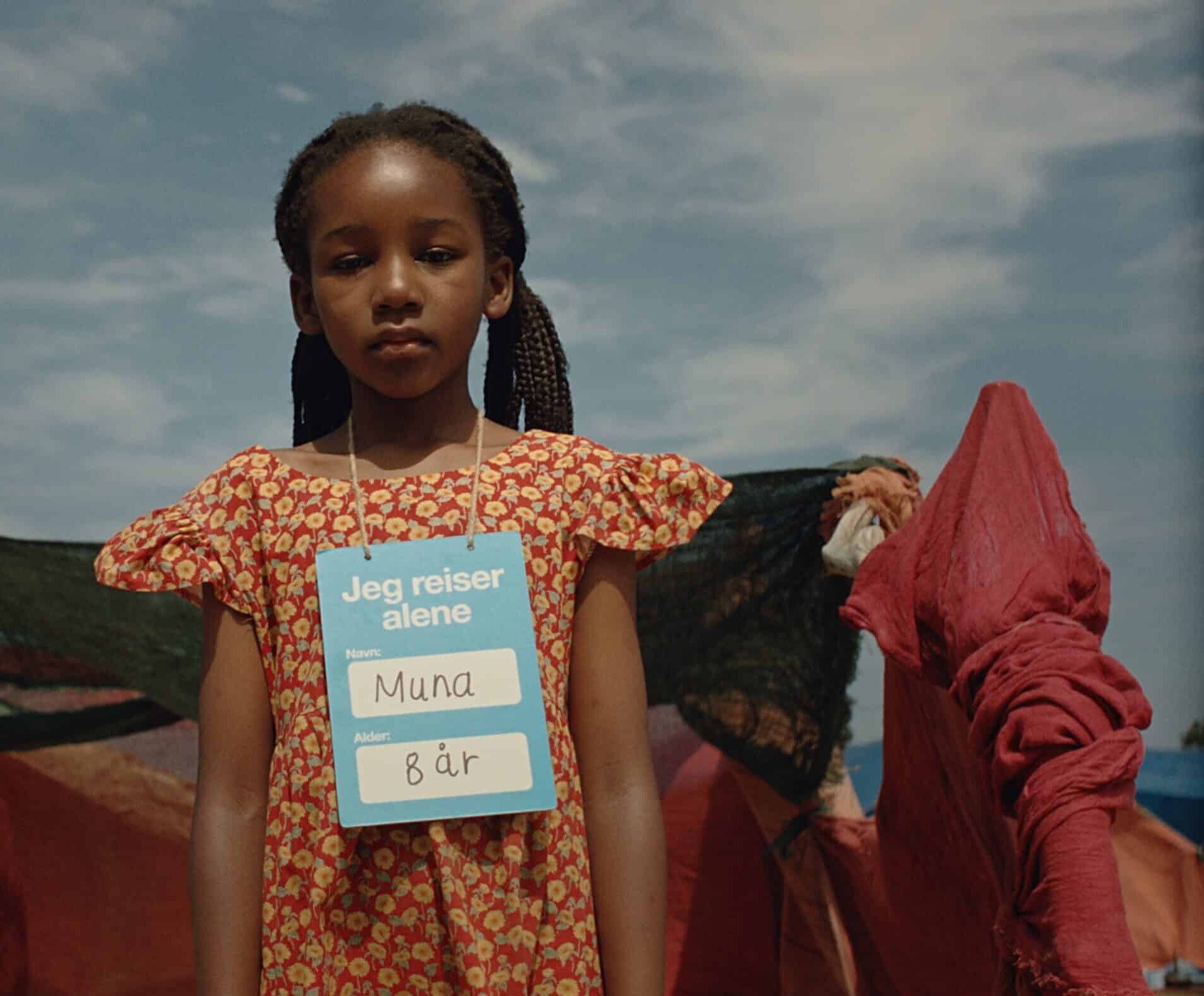 En jente på åtte år står ute med et skilt rundt halsen der det står «Jeg reiser alene». Bildet er fra kampanjefilmen til SOS-barnebyer og symboliserer barn i Sudan som vokser opp alene i kriseområder.