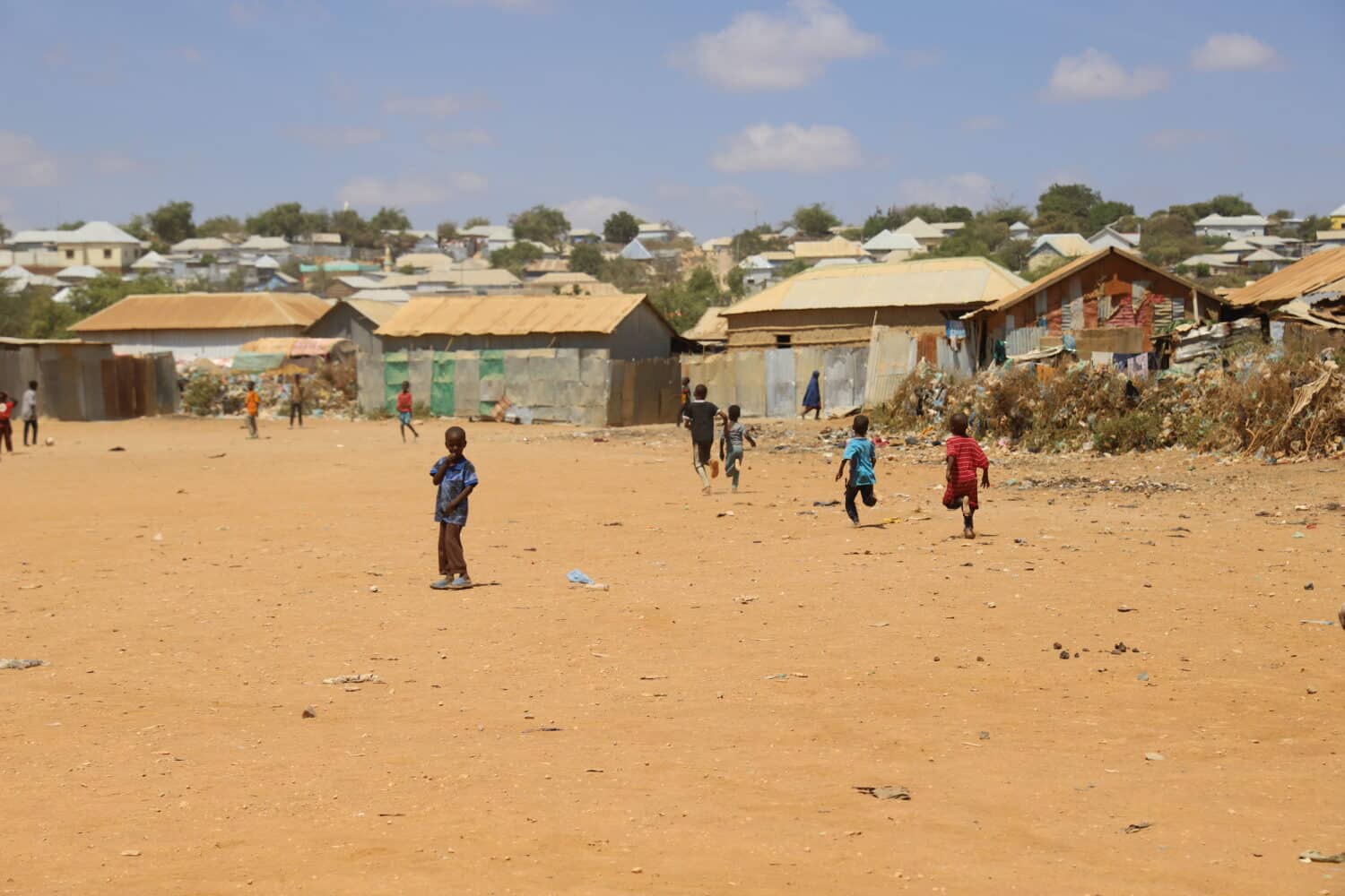 Liten gutt som ser mot kamera midt på en støvete åpen slette, med et par andre barn som løper i bakgrunnen.