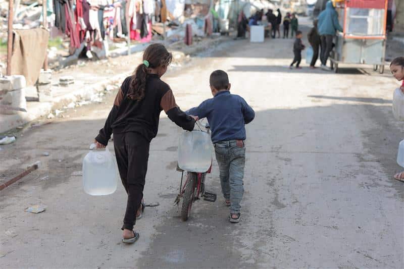 To barn som går alene med en sykkel mellom seg og bærer tunge bøtter med vann