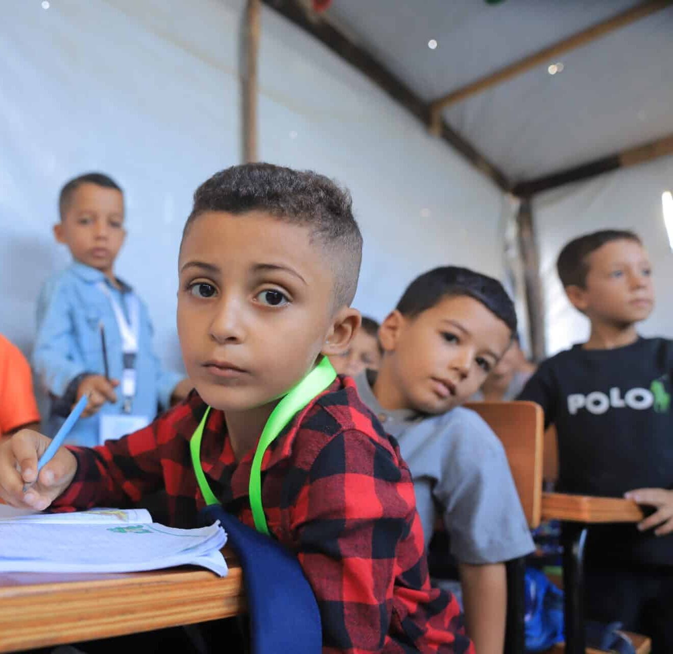 En gutt som sitter inne i et læringssenter i Gaza med penn i hånden og skrivebok foran seg og ser inn i kamera.