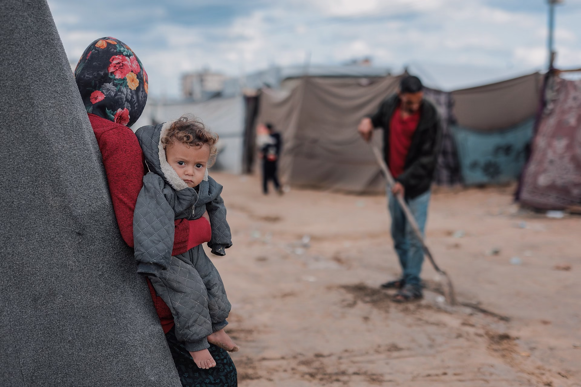 Et lite barn i en flyktningleir i Gaza. Blir holdt av en kvinne i rød genser. Har på seg en grå dress og er barbeint.