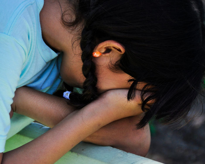 Jente fra Ecuador med mørkt hår i fletter og lyseblå t-skjorte, bøyer seg framover og skjuler ansiktet i hendene. Illustrasjonsfoto: Fernando Espinoza