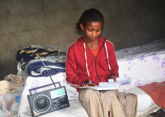 Tigist (13) sitter på senga og jobber med lekser. Ved siden av seg har hun en soldrevet radio så hun kan følge fjernundervisningen. Foto: Nigus Hailu
