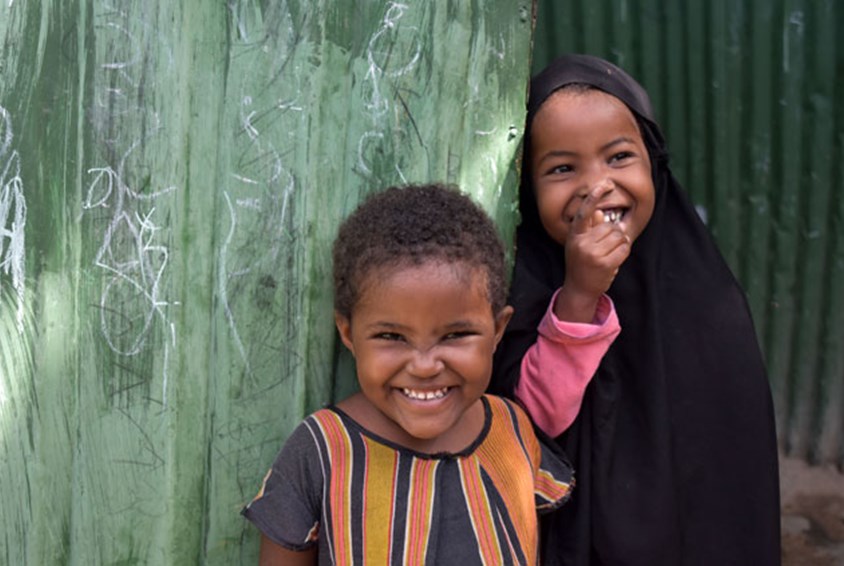 Lillesøsteren og lillebroren til Tasha står ved en vegg og smiler. Hun har på seg svart hijab og rosa genser, han har stripete, kortermet genser. Foto: Christine Stolz