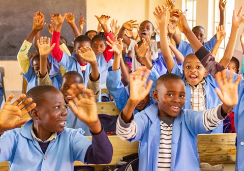 Skoleelever med lyseblå skjorter som skoleuniform sitter ved pultene og strekker armene i været. Foto: SOS-barnebyer