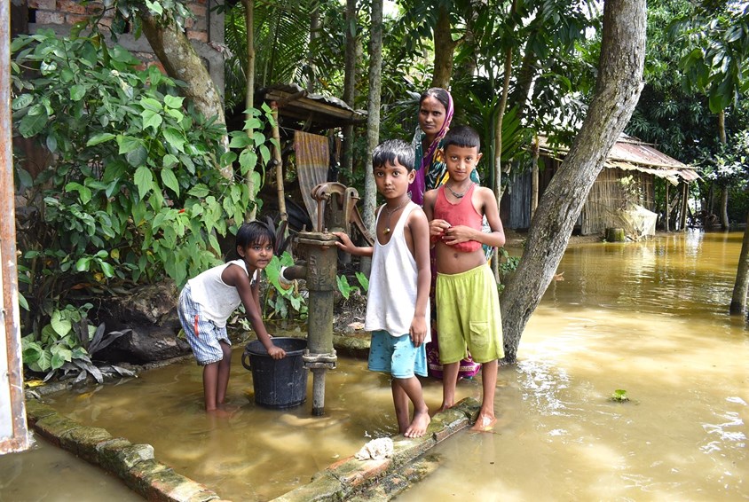 Mor og barn står i et oversvømt område i Sylhet. Foto: SOS-barnebyer