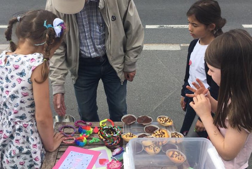 En eldre mann med beige cap og jakke handler på basar til tre barn som selger muffins og leker.