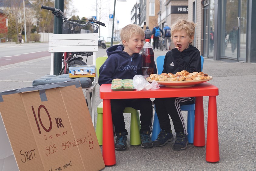 To gutter sitter ved et lite rødt bord og selger saft og kanelsnurrer til inntekt for SOS-barnebyer.                               