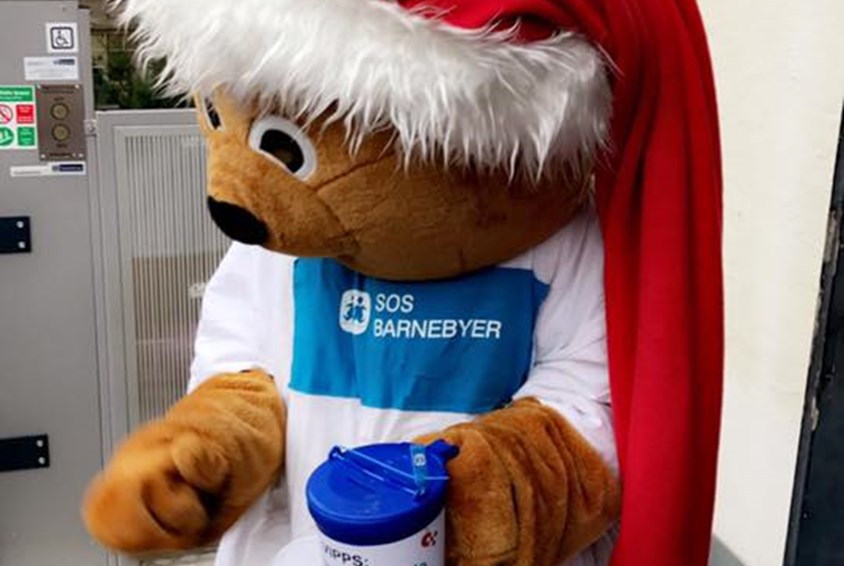 SOS-bamsen med julenisselue og hvit t-skjorte og en blå bøsse i hånda.
