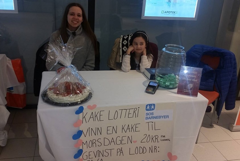 To jenter selger lodd til kakelotteri med plakat til inntekt for SOS-barnebyer foran bordet.