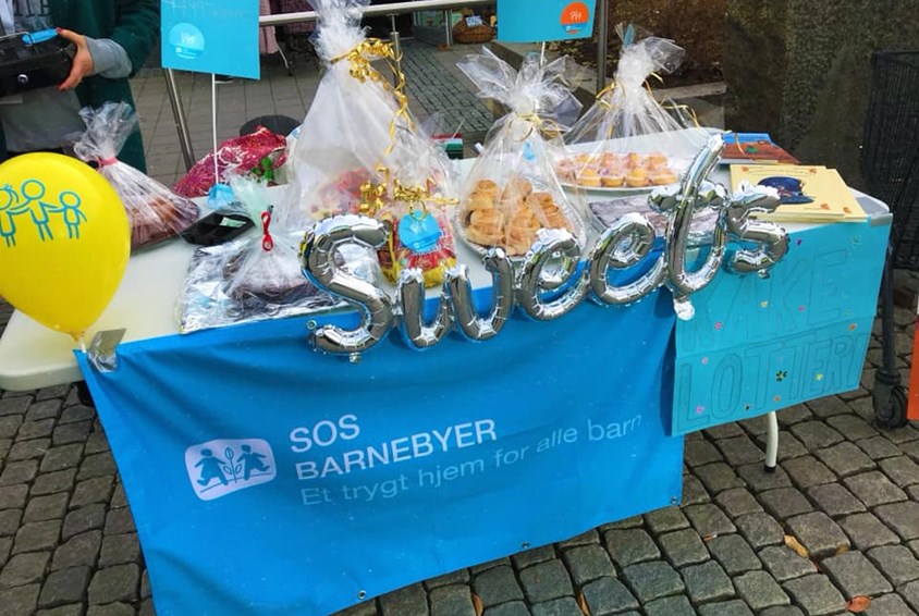 Standbord med flotte innpakkede kaker til kakelotteri. Blått SOS-banner foran bordet.