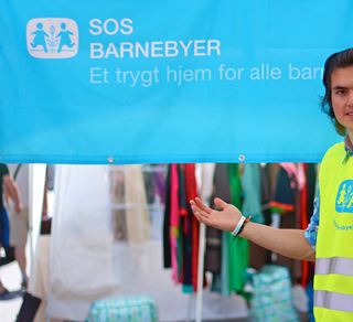 En ung gutt med gul SOS-vest står til høyre i bildet og viser med hånda det blå SOS-barnebyer banneret.