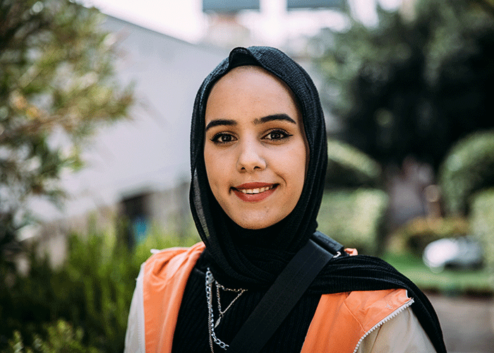 Portrett av Haneen som står ute og som smiler, hun har på seg svart genser, beige jakke og hijab. Foto: Alea Horst