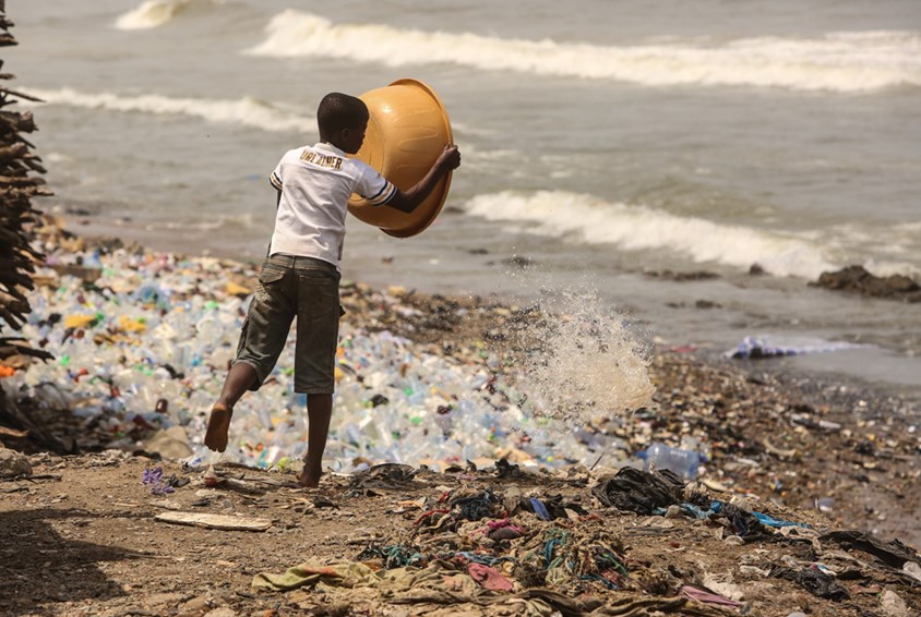 Gutt står på en strand overfylt av plastsøppel og tømmer en balje. Illustrasjonsfoto: Tom Maruko