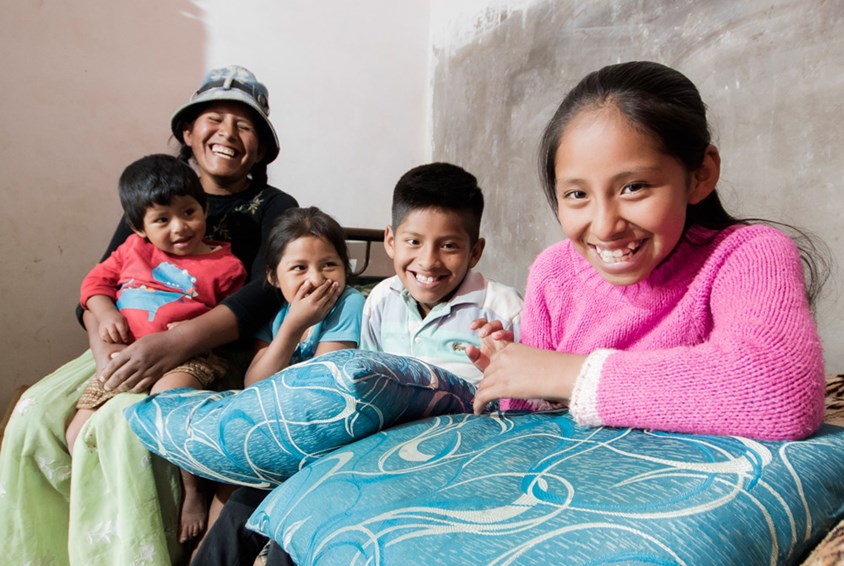 Carla* sitter i en seng sammen med moren og to søsken. Alle smiler. Foto: SOS-barnebyer