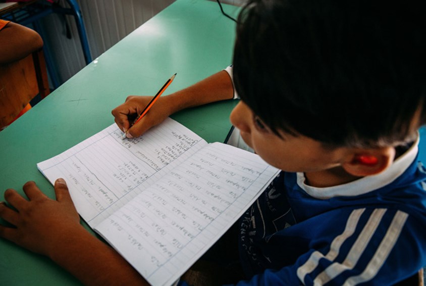 Gutt, fra flyktningleiren Kara Tepe, sitter ved en pult og skriver i ei bok. Foto: Alea Horst