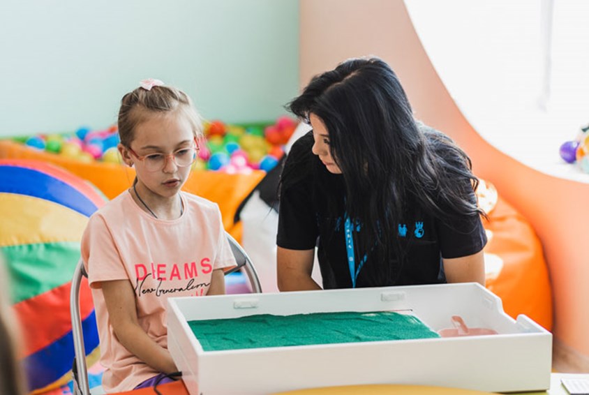 En medarbeider fra SOS-barnebyer med mørkt hår og svart t-skjorte sitter ved et bord og snakker med ei jente med blondt hår, briller og rosa t-skjorte. Foto: Ksenia Borisova
