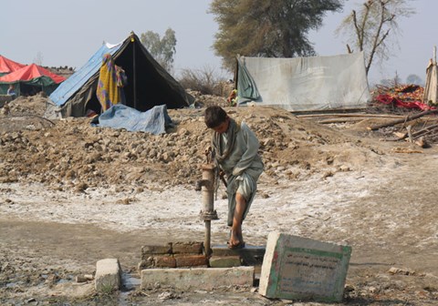 En gutt står i et tørt landskap ved ei vannpumpe, det er nødhjelpstelt i bakgrunnen. Foto: Muhammad Ali