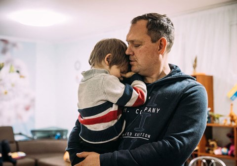 En SOS-fosterfar, med mørkeblondt kort hår og blå hettegenser, holder en liten gutt i armene, han har på seg stripete genser og mørk bukse. Foto: Alea Horst