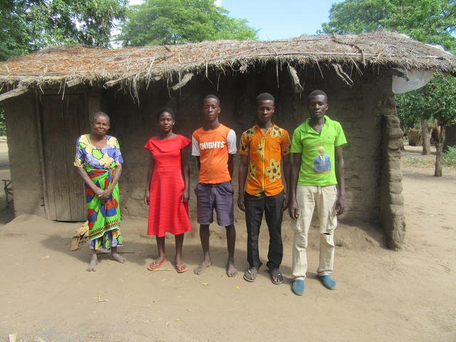 Kozani med brødrene og bestemoren sin. De står utenfor huset sitt, som er av leire og har stråtak. 